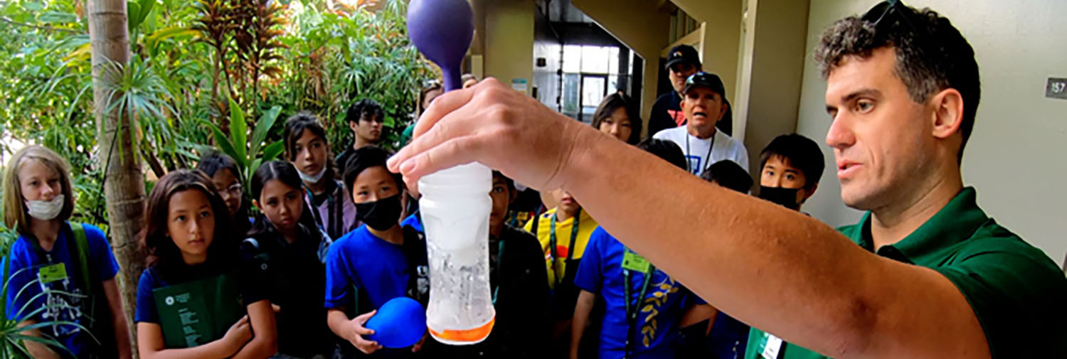Adult Male Instructor Showing Shows A Group Of Intermediate Students A Balloon Attached To A Plastic Bottle